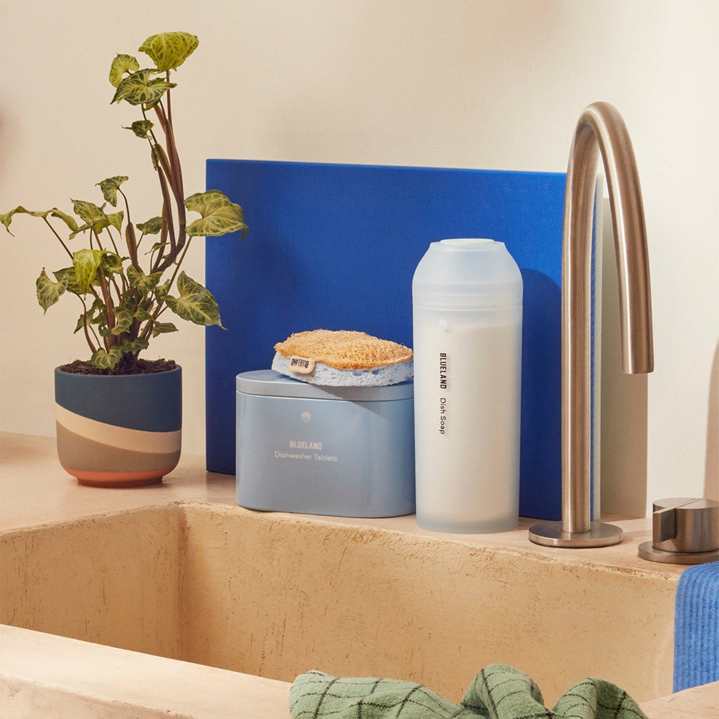 A kitchen sink area features waste reduction items: a beige countertop, modern silver faucet, striped pot plant, blue dishwasher tablet container, sponge, and a 1 Year of Dish Soap bottle. A green cloth hangs from the sink.