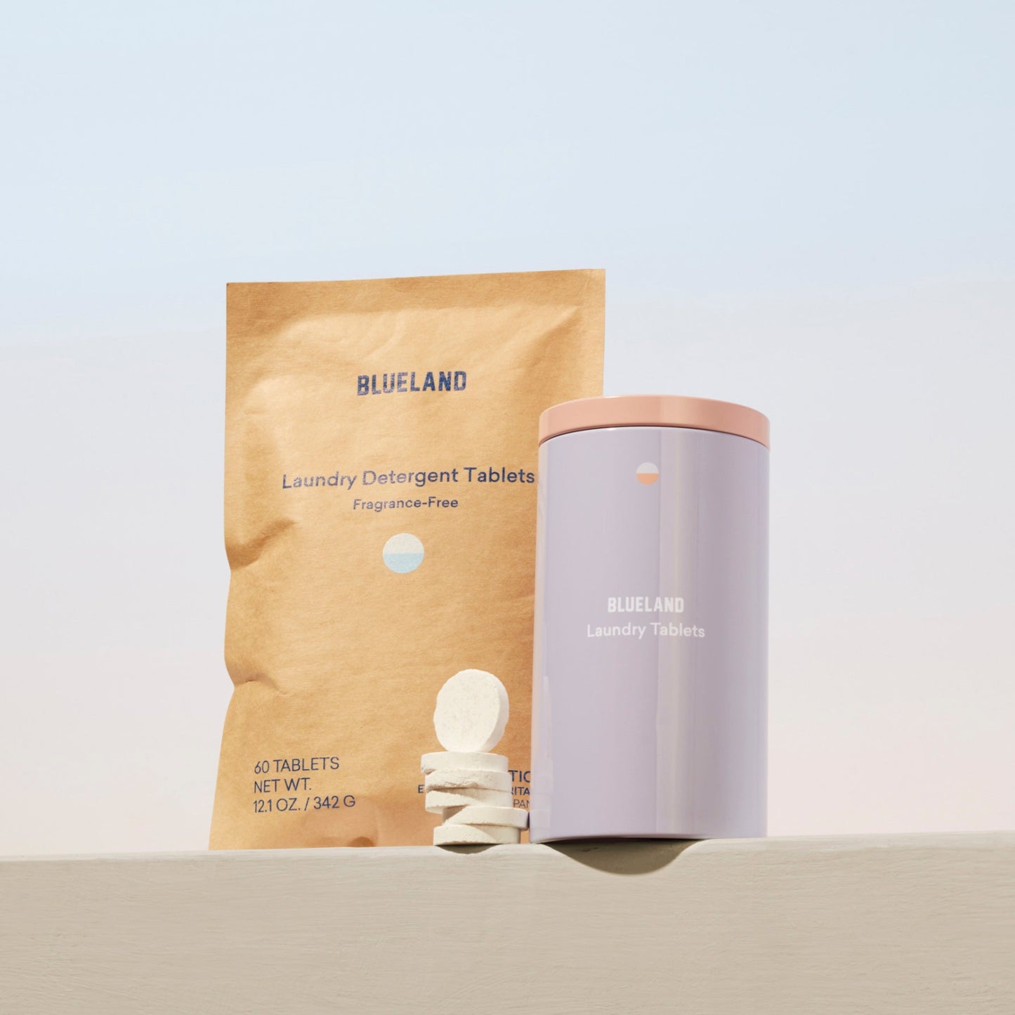 A package of the Laundry Detergent Starter Set by Blueland, known for its eco-friendly, plastic-free tablets, is beside a lilac container with a pink lid. Several white tablets are stacked in front on brown packaging with blue text against a light blue and beige backdrop.