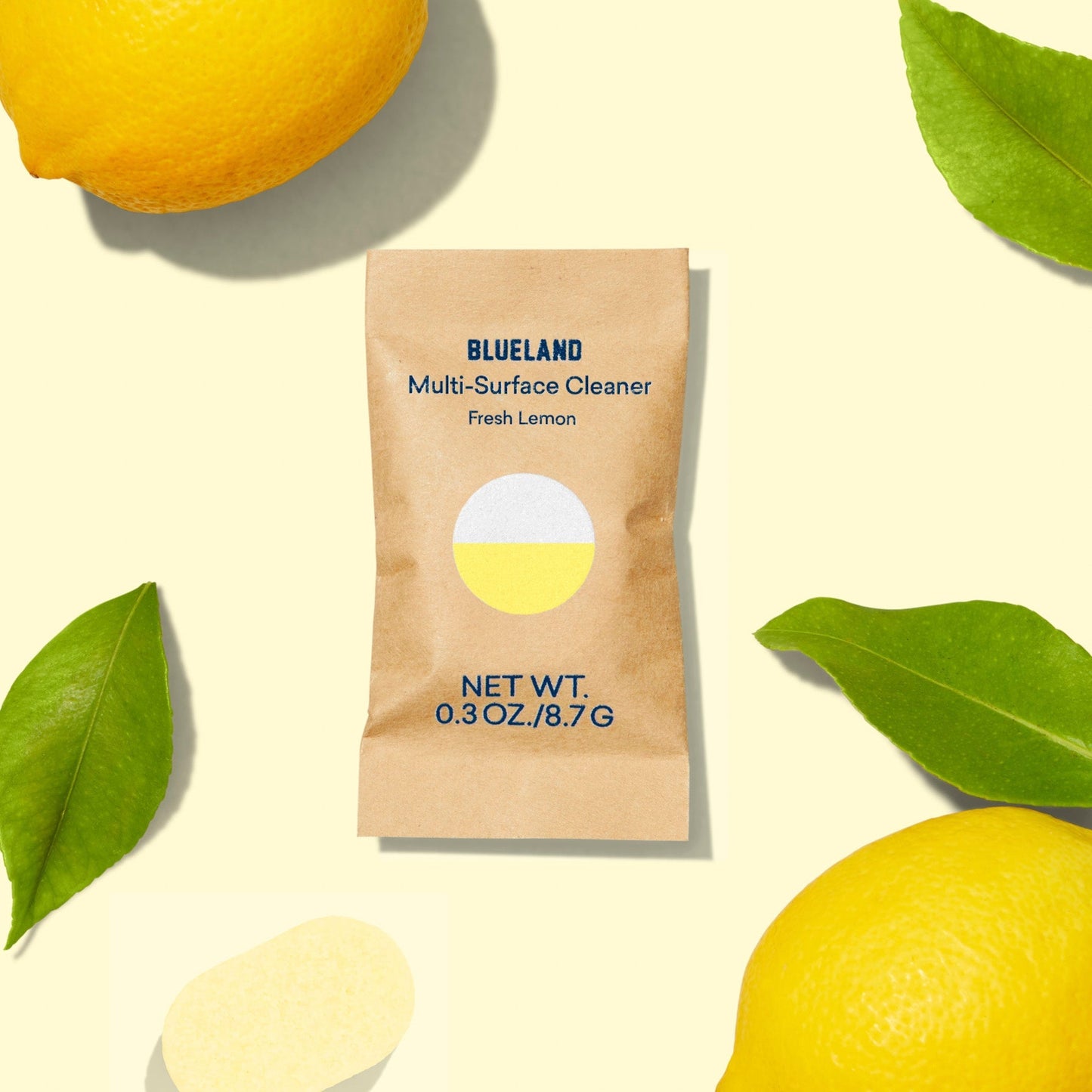 A packet of Multi-Surface Cleaner in Fresh Lemon scent is centered on a light yellow background, surrounded by fresh lemons and green leaves. The packet shows a net weight of 0.3 oz/8.7 g.