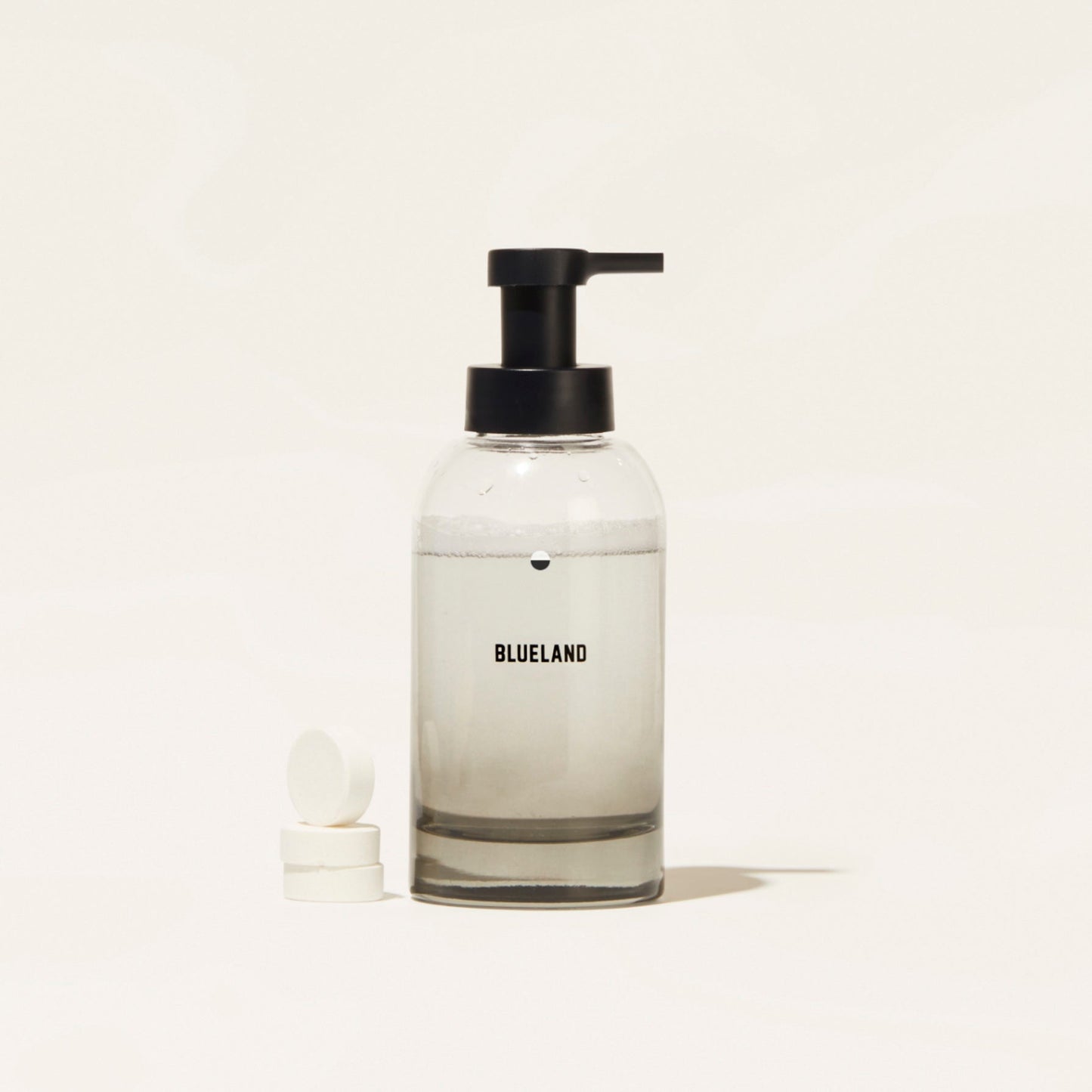 A Blueland Hand Soap Starter Set (Bundle) featuring a clear pump bottle filled with foaming soap made from clean ingredients stands beside two white tablets on a plain, light background.