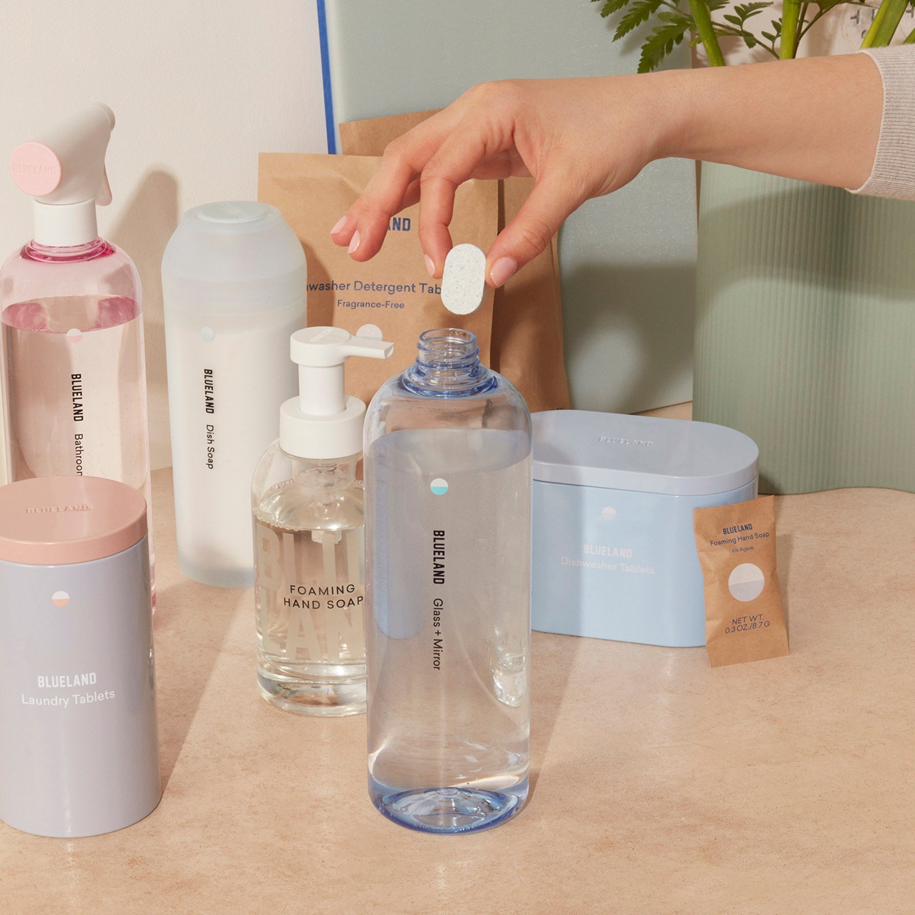 A hand inserts a refill tablet into a labeled Glass + Mirror Cleaner bottle. Nearby, other Blueland items, such as plant-powered cleaners in pink bottles, foaming hand soap, and detergent packs are placed on a beige surface with a leafy plant in the background.