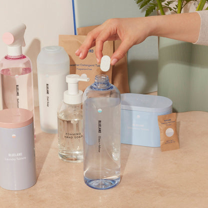 A hand inserts a refill tablet into a labeled Glass + Mirror Cleaner bottle. Nearby, other Blueland items, such as plant-powered cleaners in pink bottles, foaming hand soap, and detergent packs are placed on a beige surface with a leafy plant in the background.