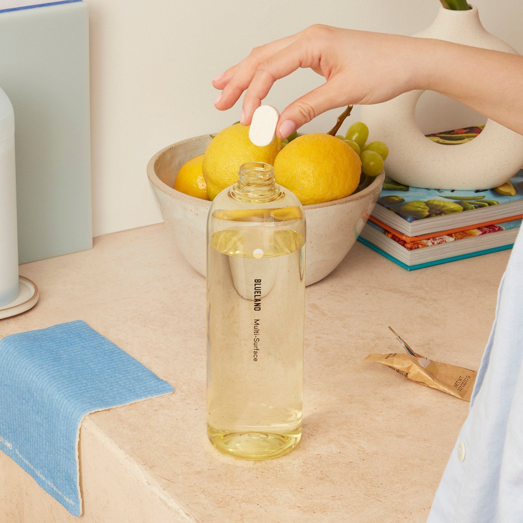 A person drops a Multi-Surface Cleaner Tablet into a clear bottle labeled Blueland on a countertop. Nearby, there is a bowl of lemons and grapes, some books, a closed towel, and a small piece of paper.