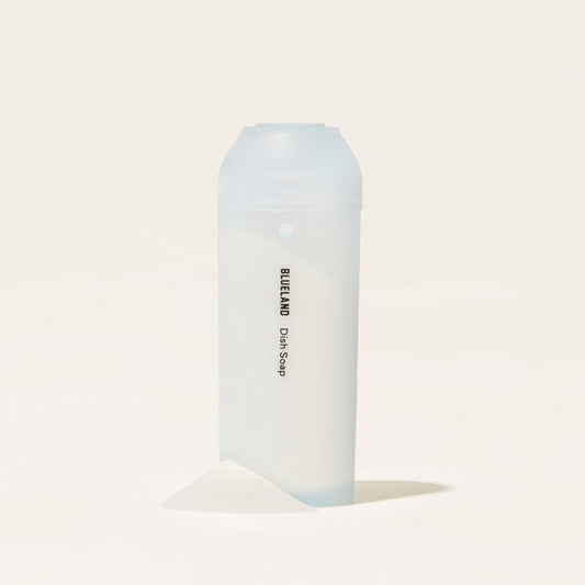 A translucent Dish Soap Starter Set bottle stands upright on a white surface, featuring its plant-based formula. A small powder mound is placed in front against a plain background, highlighting an eco-friendly clean and waste reduction.