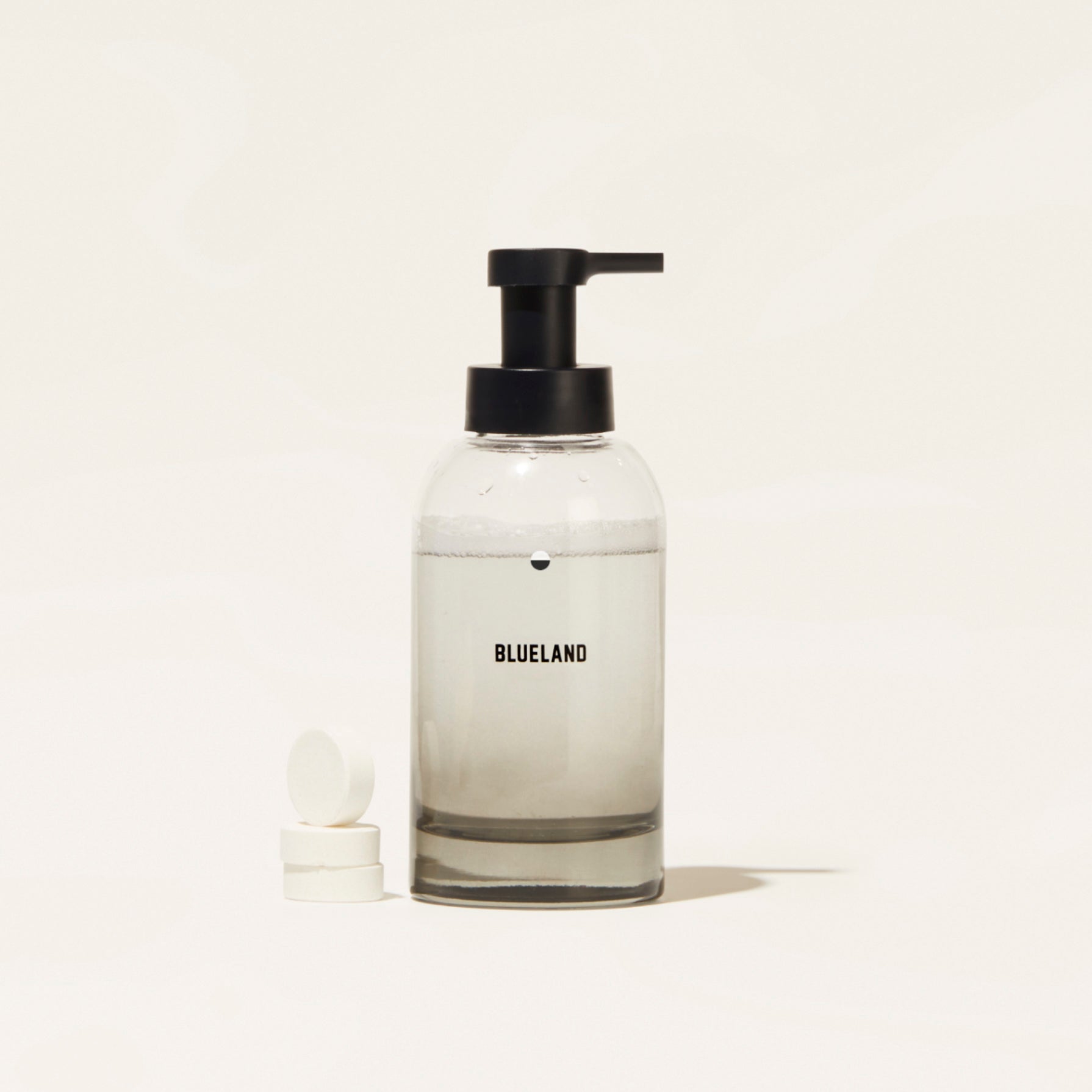 A Blueland Hand Soap Starter Set (Bundle) featuring a clear pump bottle filled with foaming soap made from clean ingredients stands beside two white tablets on a plain, light background.
