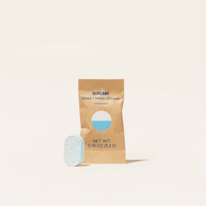 A small blue tablet stands beside a brown packet labeled Glass + Mirror Cleaner Tablet Refills, Unscented with a blue circle, net weight 0.18 oz (5.2 g). This plant-powered cleaner uses refill tablets and is set against an off-white background.